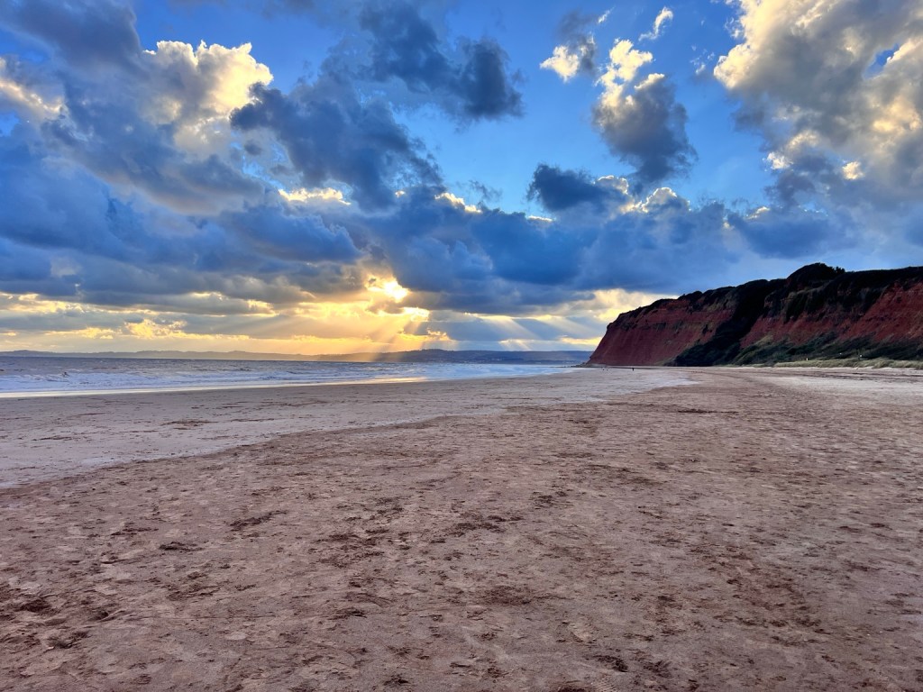 Golden sunlight streams through clouds over a broad sandy beach and the red cliffs at Devon Cliffs, Exmouth