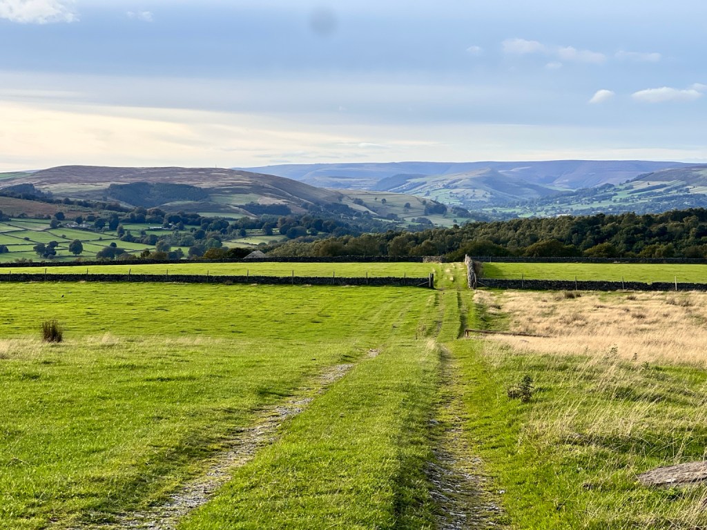 A grassy track between fields overlooking green valleys and distant Peak District hills.